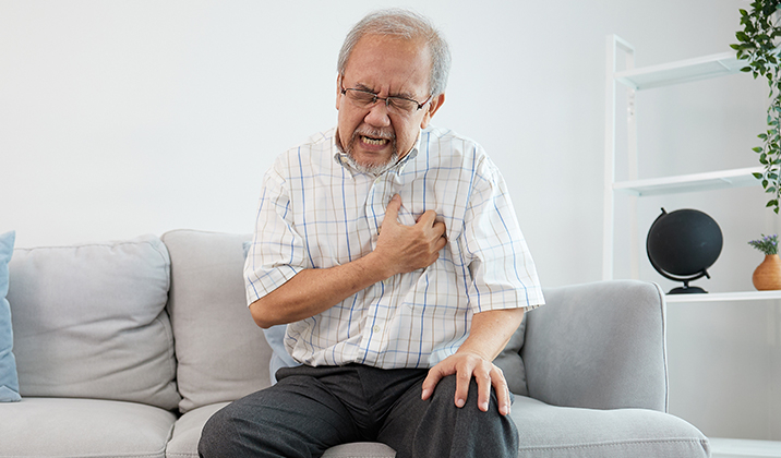 Elderly Asian man on a grey sofa, clutching his chest in distress, indicative of a heart attack.